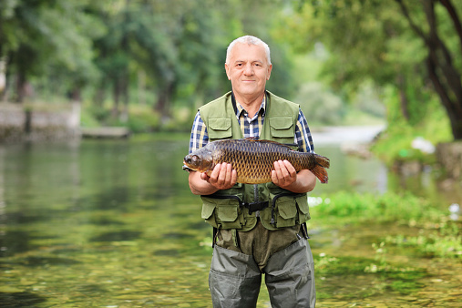 Mature fisherman standing in river and holding a fish outdoors