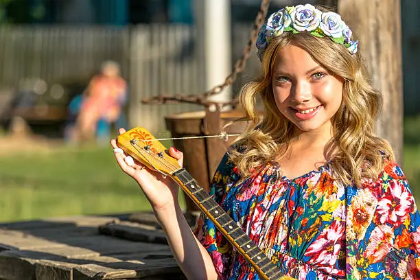 Beautiful teen blonde girl standing with balalaika at sunny summer day in countryside