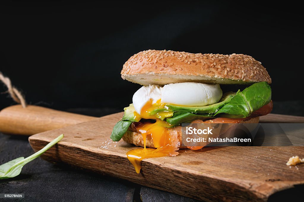 Bagel with salmon and egg Bagel with salted salmon, spinach, avocado and soft boiled egg with liquid yolk on wooden chopping board over old wood black background. Avocado Stock Photo