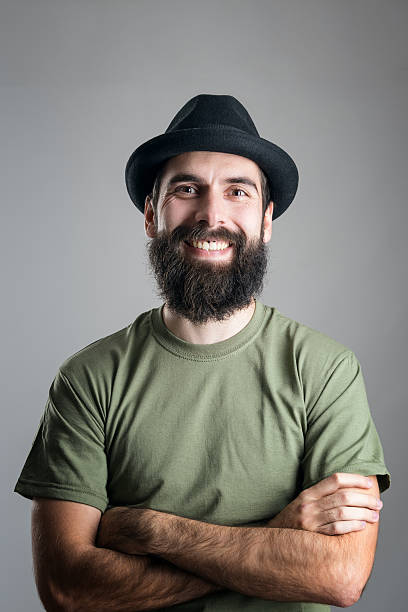 Bearded hipster wearing hat with upper lips piercing friendly laughing Bearded hipster wearing hat with upper lips piercing friendly laughing at camera. Headshot portrait over gray studio background with vignette. people laughing hard stock pictures, royalty-free photos & images