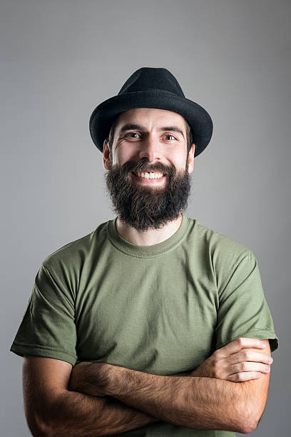 Bearded hipster wearing hat with upper lips piercing friendly laughing Bearded hipster wearing hat with upper lips piercing friendly laughing at camera. Headshot portrait over gray studio background with vignette. man laughing hard stock pictures, royalty-free photos & images