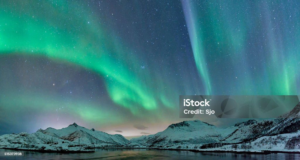 Northern Lights over the Lofoten Islands in Norway Northern Lights, polar light or Aurora Borealis in the night sky over the Lofoten islands in Northern Norway. Wide panoramic image with snow covered mountains and a lake in the foreground. Aurora Borealis Stock Photo