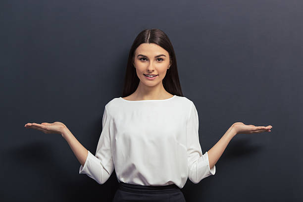 Beautiful young student Beautiful young student in classic elegant clothes is is smiling, looking at camera and keeping hands spread showing balance, standing against blackboard blouse stock pictures, royalty-free photos & images