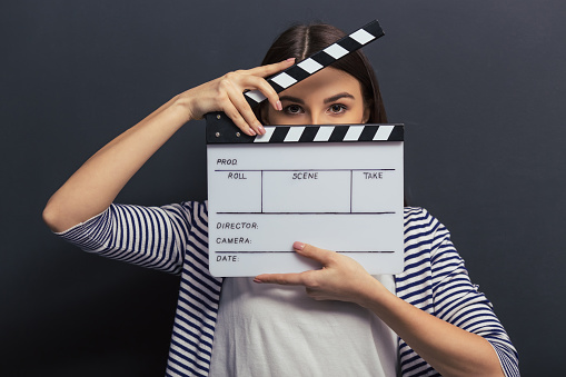 Beautiful young girl in casual clothes is covering her face with a clapperboard and looking at camera, standing against blackboard