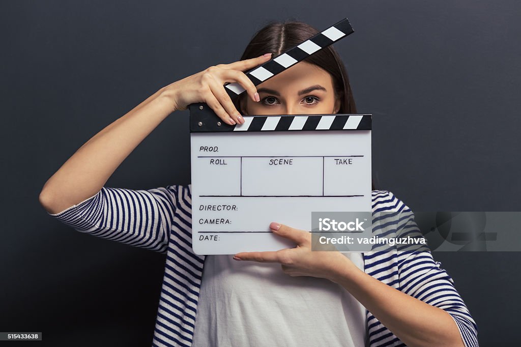 Handsome girl with clapperboard Beautiful young girl in casual clothes is covering her face with a clapperboard and looking at camera, standing against blackboard Movie Stock Photo