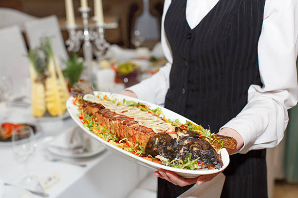 waitress in a white blouse and a black uniform holding a white plate with baked fish in a restaurant