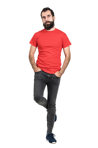Bearded hipster in red t-shirt standing and balancing on one leg looking away. Full body length portrait isolated over white studio background.
