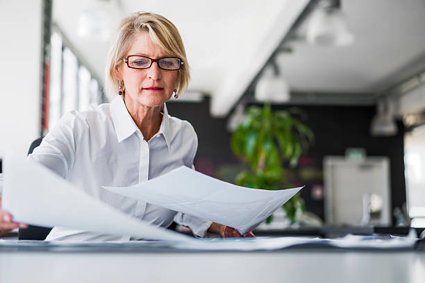 Businesswoman examining documents at desk A photo of mature businesswoman examining documents at desk. Concentrated professional is analysing papers in office. Executive is in formals. analyzing document stock pictures, royalty-free photos & images