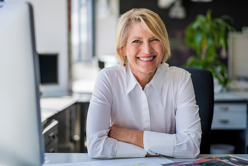 A photo of happy mature businesswoman sitting at desk. Portrait of female professional is in office. Smiling executive is in formals at workplace.