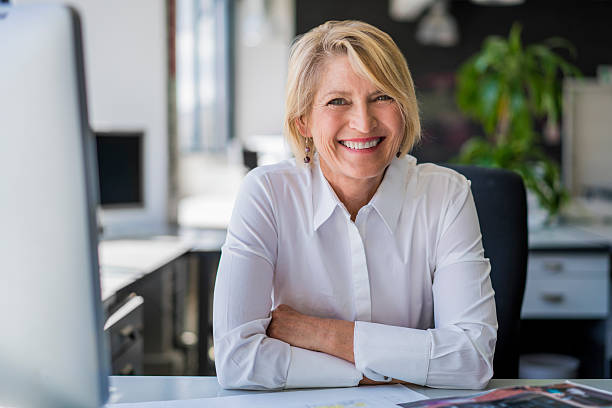 Happy mature businesswoman sitting at desk A photo of happy mature businesswoman sitting at desk. Portrait of female professional is in office. Smiling executive is in formals at workplace. older blonde woman stock pictures, royalty-free photos & images