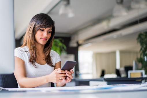 A photo of beautiful businesswoman using smart phone at desk in office. Female executive is in formalwear. Professional is text messaging on mobile phone at workplace.