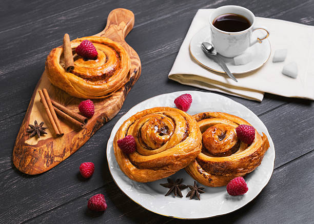 Round twisted Denish Buns Round twisted Denish Buns with berries raspberries, syrup, white cup of espresso coffee, sugar cubes, cinnamon sticks, star anise, cutting board made of wood olive on a black wooden background denish stock pictures, royalty-free photos & images