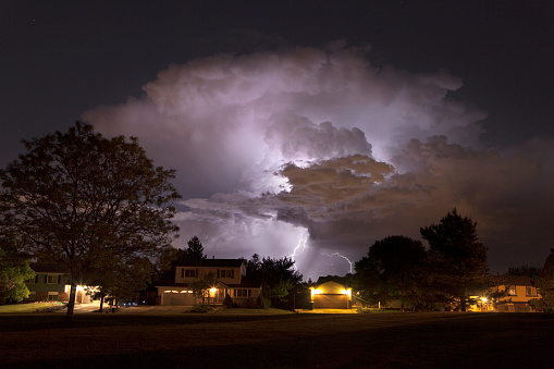 At night as rain falls and stars shine in the distance, lightning bolts strike over southwest suburban Denver homes and trees and the green grass of a local Colorado park. 