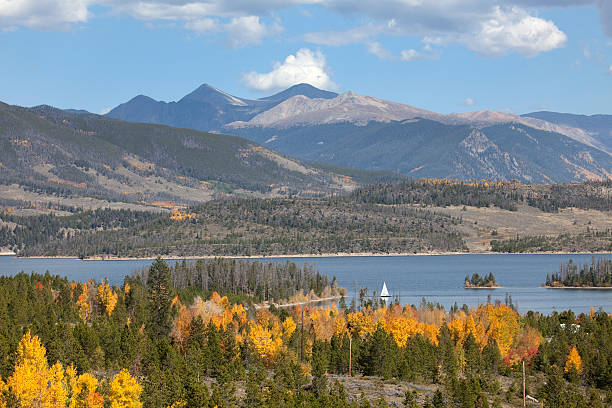 With yellow aspen and pine trees covering the hillsides, a sailboat cruises the waters of Lake Dillon or Dillon Reservoir in Summit County. The 14,000+ foot mountains of Grays (on the right) and Torreys Peaks (pyramid shaped peak in the middle rise into a blue autumn sky.