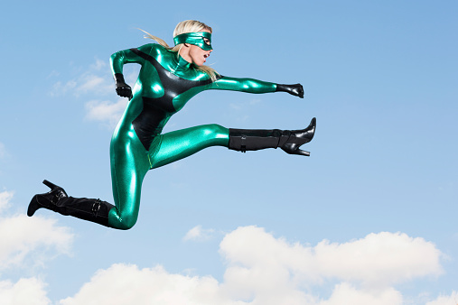 A beautiful young caucasian woman super hero flys through the air to deliver a punch kick. Mid air with sky and clouds in the background.