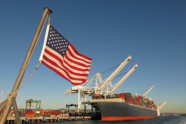 American flag US port container ship symbols economy industry pride An American flag waves in the foreground at this US port, where a cargo ship loaded with containers is berthed beneath giant cranes on a clear blue sky day. The image symbolizes concepts like Made in America, Made in the USA, industry, global economy, pride, strength, power, patriotism, trade, manufacturing, and the idea of getting back to work. quayside photos stock pictures, royalty-free photos & images