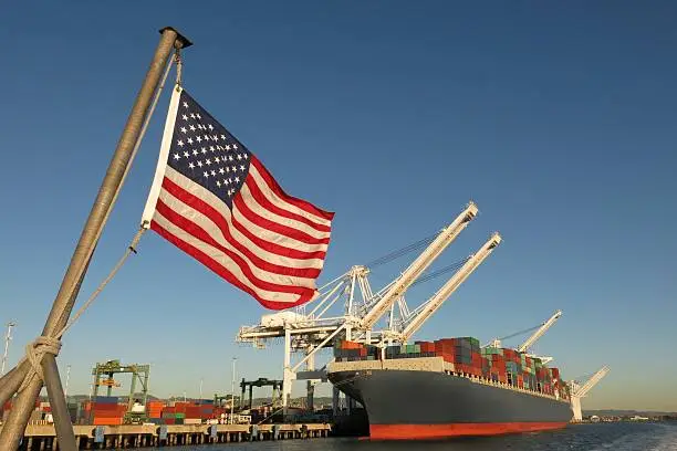 An American flag waves in the foreground at this US port, where a cargo ship loaded with containers is berthed beneath giant cranes on a clear blue sky day. The image symbolizes concepts like Made in America, Made in the USA, industry, global economy, pride, strength, power, patriotism, trade, manufacturing, and the idea of getting back to work.