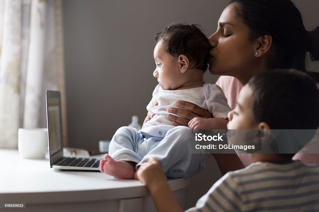 Watching cartoons on mummy's laptop Young mother sitting with her two sons at the dining table. They are watching cartoons on a laptop and the mother is kissing her baby son on the head. Pregnant Stock Photo