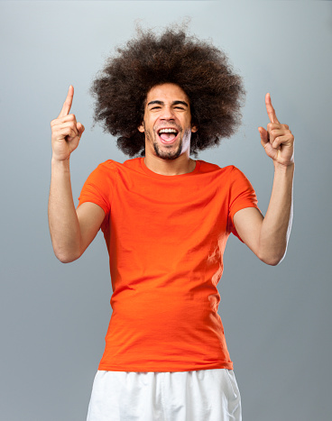 three quarter length shot of a screaming and shouting man, making number one sign with big afro hair in orange T-shirt fan of the Dutch soccer team, looking at the camera on gray background
