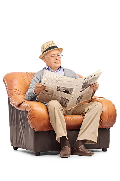 Vertical shot of a relaxed senior gentleman reading a newspaper seated on an armchair isolated on white background