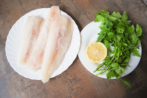 Frozen Alaska Pollock fillet with lemon and parsley on white dish. Preparation for cooking.