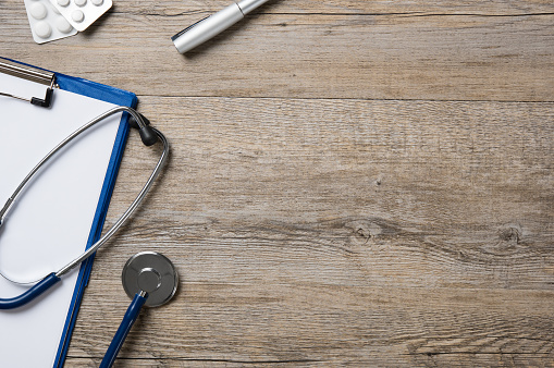 Top view of medical blue clipboard and stethoscope on wooden desk with copy space. Wooden doctor desk with stethoscope, medicine, clipboard and pills. Healthcare background concept.