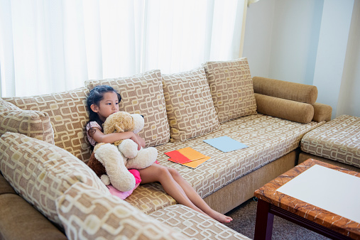 This is a horizontal, color photograph of a 6 year old Thai girl. She sits inside on a couch holding a stuffed animal while she watches tv.