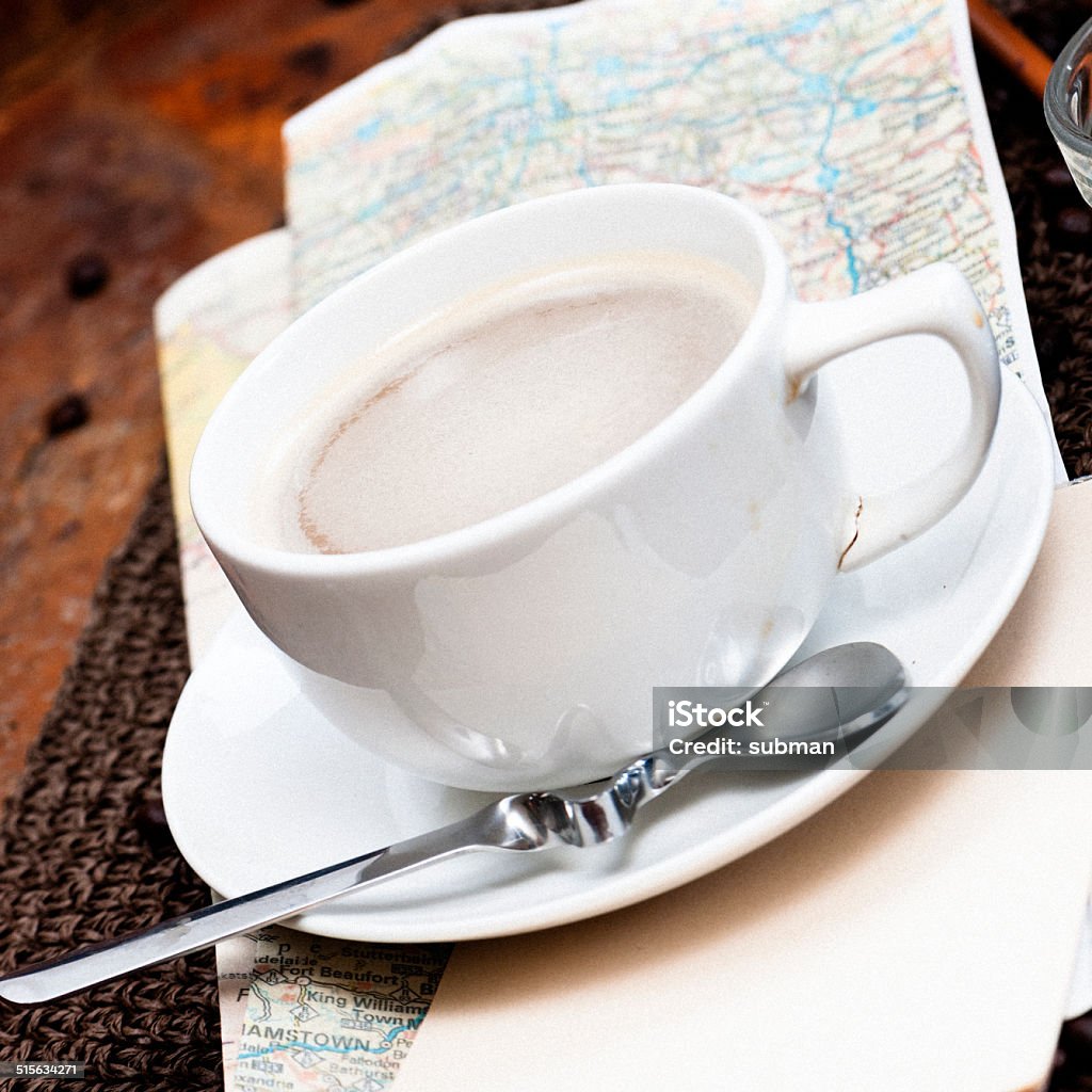 Angled close-up of a cup of coffee sitting on a roadmap. Coffee in white coffee cup & saucer placed on a road map on wooden table. Brown Stock Photo