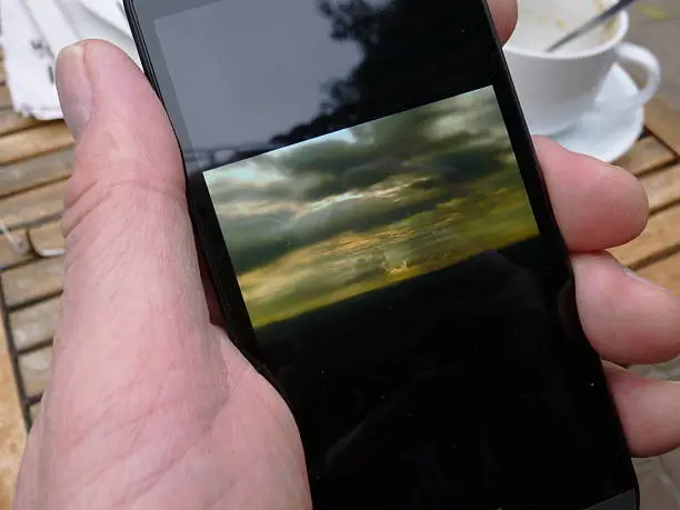 Hand of a male person that holds a Smartphone with a landscape-photo on it