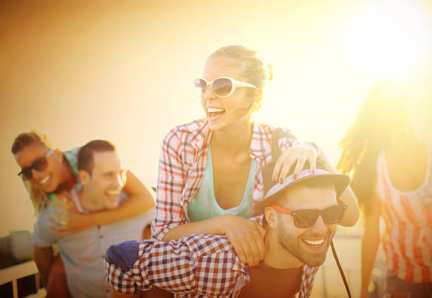 Group of mid 20's people walking the street and having fun. There are two guys and three girls, two of them wearing sunglasses. Guys are piggybacking their girls. All laughing and enjoying summer. 