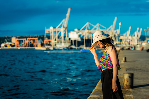 Happy young Chinese tourist/student standing by the Adriatic Sea, smiling and looking at the water surface. She has long dark hair, she is wearing a white straw hat with polka dot ribbon, a sleeveless shirt with stripe pattern and a b long black skirt.  Big shipyard with cranes blurred in the background, Koper, Slovenia, Adriatic Sea, Europe. Deep blue colour. Nikon D800, full frame, XXXL.