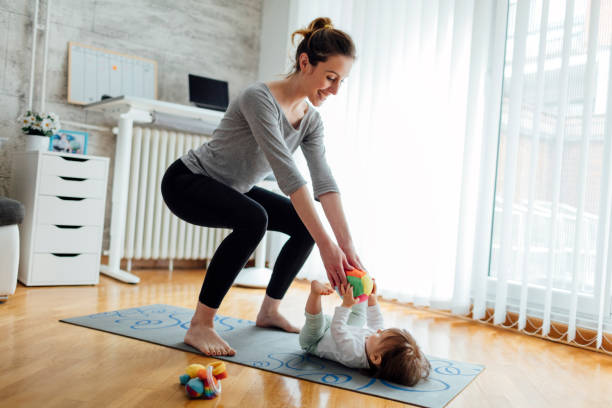 Mother Exercise With Her Baby At Home Shot of an attractive young woman with her baby girl while doing exercise. Woman working squats and baby lying on exercise mat. They exercise in a living room. Mother playing with her baby while exercise. baby squatting stock pictures, royalty-free photos & images