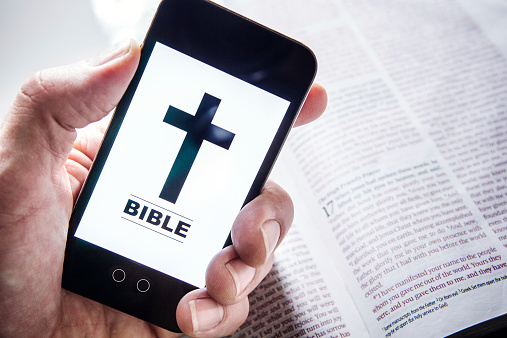 ILLUSTRATION AND PHONE BUTTONS ARE MY OWN DESIGN.  A mans hand holds up a smart phone with a digital Bible application open on the screen, an actual physical Bible laying open in the background.  Horizontal image with copy space.