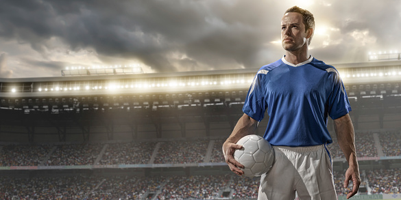 A front view portrait image of soccer player from waist up holding football and looking off camera. Football player is standing in a generic floodlit soccer stadium under a dramatic cloudy evening sky at sunset. Soccer player  is wearing a generic unbranded blue soccer shirt. All stadium advertising is fake. 