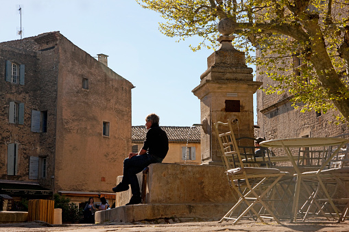 Gordes, Provence, France -- April 9, 2014: Man dressed in black is sitting on the rim of the fountain at the central square of Gordes, Provence, France. Restraints and small shops are located at the first floor of the houses, accidental people are walking on the street.
