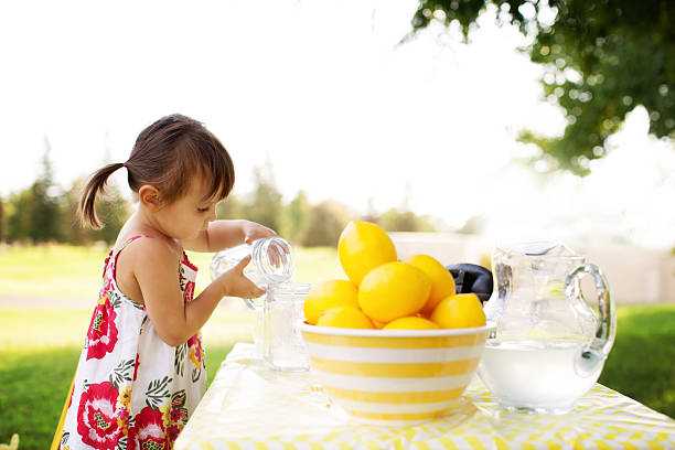 Little Girl At The Lemonade Stand Little Girl At The Lemonade Stand Transferring Lemonade From One Cup to Another Outdoor in A Summer Day. images of lemonade stands stock pictures, royalty-free photos & images