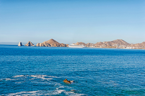 This view is looking across the bay in Cabo San Lucas taking in the iconic signature rocks welcoming visitors with a cruise ship in port. and the cityscape on the hillside.