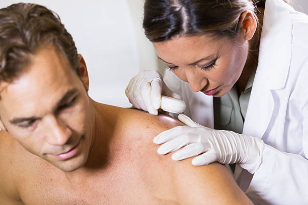 Dermatologist examining patient's skin for signs of cancer Female dermatologist (30s, mixed race, Asian / Caucasian) examining male patient (30s) with dermascope, looking for signs of skin cancer.  Focus on woman. dermatology stock pictures, royalty-free photos & images