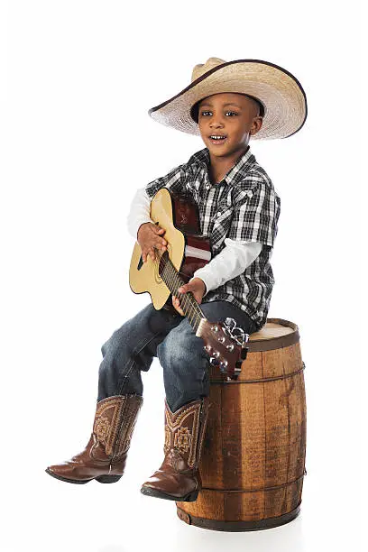 A handsome young African boy happily singing and playing his guitar as he sits on an old barrel.  On a white background.