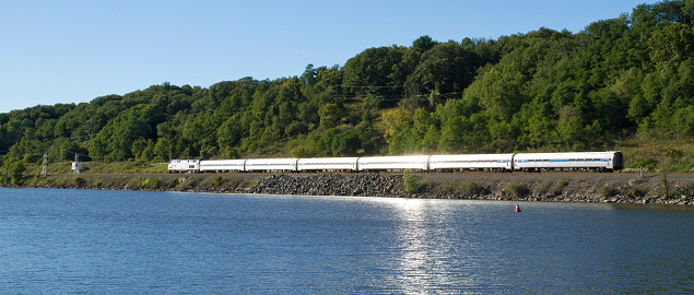 Amsterdam, NY, US - September 14, 2014: Amtrak passenger train along Mohawk river in upstate NY. Sun reflects off stainless steel cars on water. These are CSX tracks on the old New York Centrals water level route from New York City to Chicago.