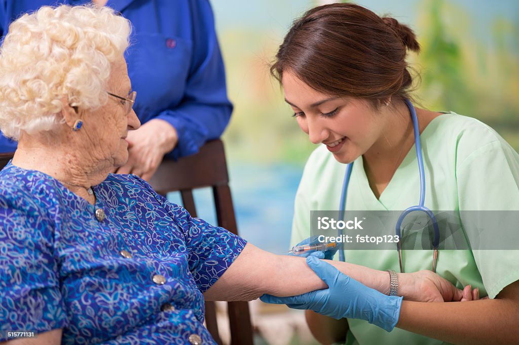 Home healthcare nurse giving injection to elderly woman. Latin descent female nurse or doctor gives vaccine or flu shot to a senior woman patient.  Home or clinic setting.  Her daughter looks on.  Woman is over 100 years old!   Nursing Home Stock Photo