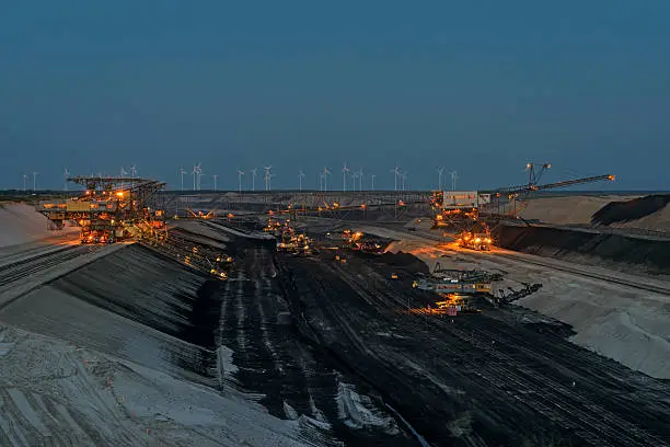 Cottbus Nord open pit at dusk. Overburden conveyor bridge with two bucket dredgers in action. The floor will removed and transported left to right across the open pit mining. The uncovered coal is dredged from coal excavators. Brown coal is transported by trains. In the background, the power plants of the future - wind turbines. Mining Cottbus Nord, Brandenburg, Germany