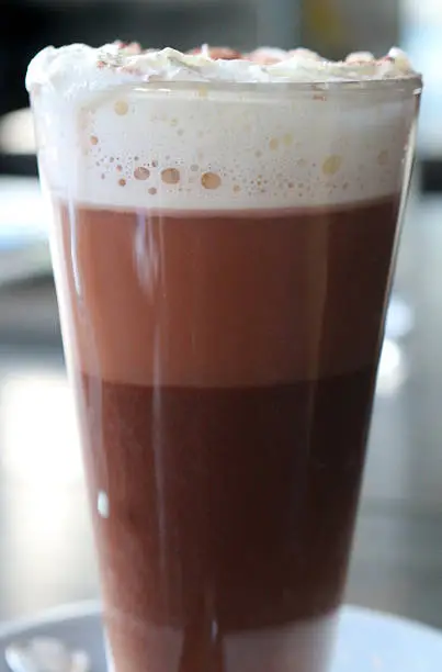 Photo showing a close-up view of a glass mug of hot chocolate topped off with whipped cream. On closer inspection it is possible to see the separating layers of cocoa, milk and cream.