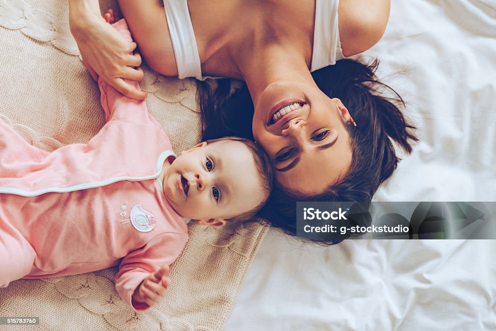 Pure happiness. Top view of cheerful beautiful young woman looking at camera with smile while lying in bed with her baby girl Mother Stock Photo