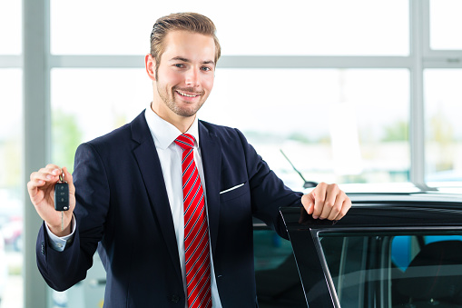 Seller or car salesman in car dealership with key presenting his new and used cars in the showroom