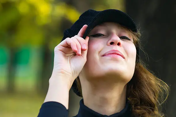 Portrait of an attractive beautiful pretty cute young caucasian cheerful smiling woman (girl, female, person, model) dressed in a black peak cap, holding it by hand and looking stright forward at you.