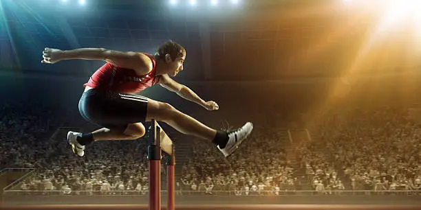 :biggrin:A professional male athlete jumping over a hurdle during a race. The action takes place in a outdoor . stadium full of spectators. The athlete is wearing generic athletics kit. Low angle image.