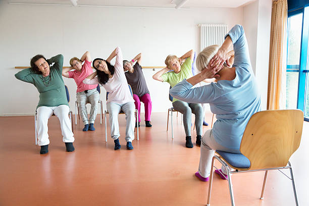 teacher and active senior women yoga class on chairs senior women exercising yoga and pilates sitting on chairs, following the instruction of their teacer chair stock pictures, royalty-free photos & images