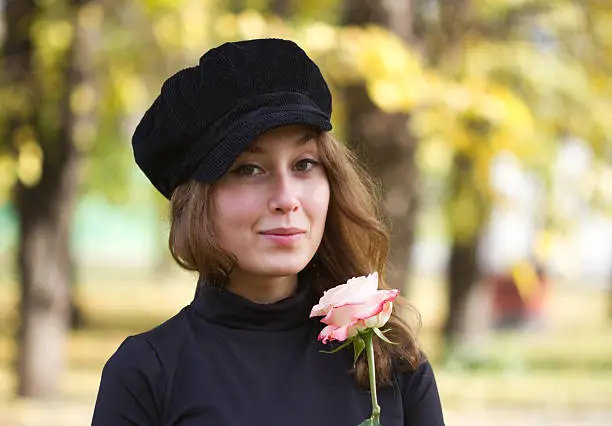 Portrait of an attractive beautiful pretty cute young caucasian cheerful smiling successful woman (girl, female, person, model) dressed in a black peak cap looking at you and holding a flower (pink rose) in her hands. Autumn park and yellow foliage serves as a blurred background.