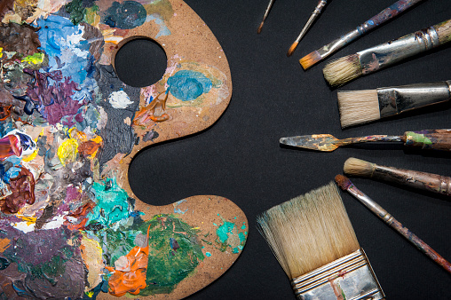 Abstract view of some used painting utensils such as a color mixing palette, a few brushes in a variety of strokes and a palette knife, all covered in multi colored paint.